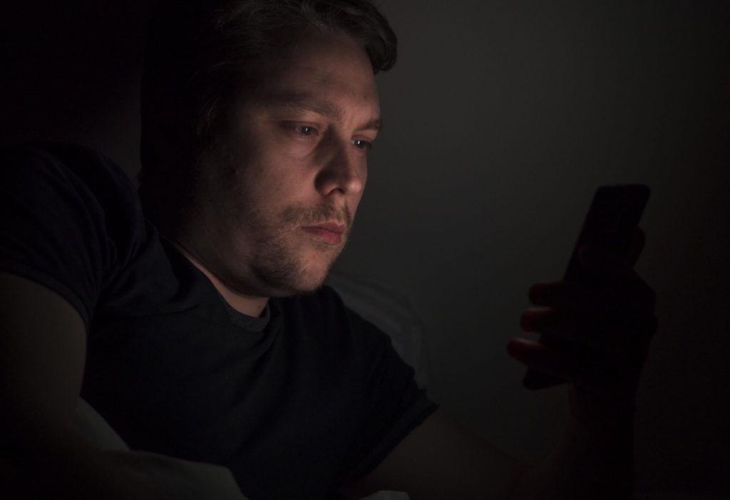 Homem deitado no escuro à noite com o rosto iluminado pela luz do celular, representando o uso compulsivo e o vício em dopamina e pornografia